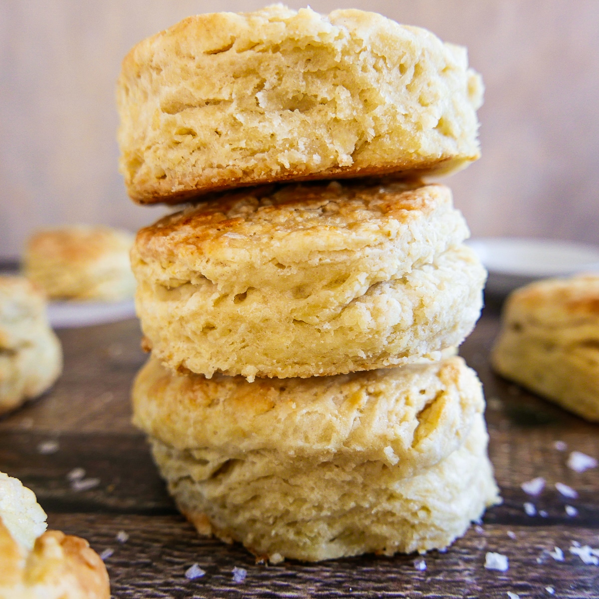 Three biscuits stacked on a wooden table with more biscuits in background.