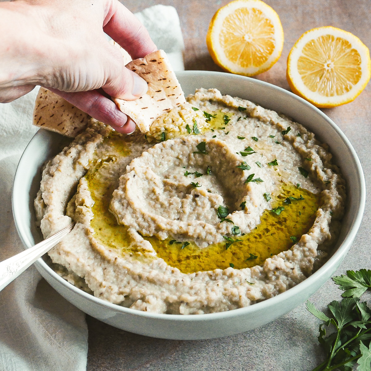 White hand dipping pita into bowl of eggplant dip with a spoon.