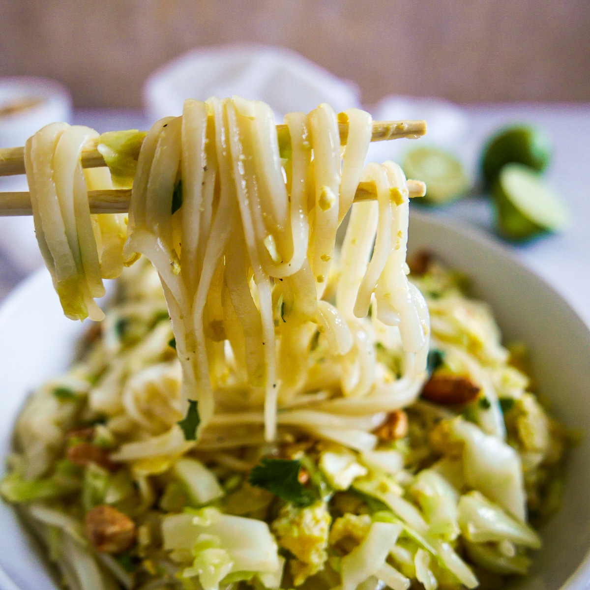 chopsticks-with-noodles.jpg Chopsticks lifting up noodles from bowl of pad thai with limes in the background.