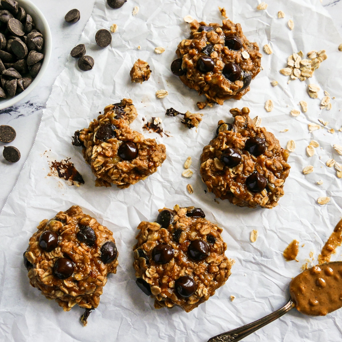 Five peanut butter banana cookies arranged on parchment with a spoon of peanut butter.