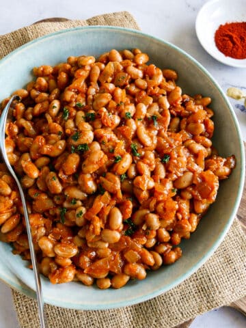 Bowl of healthy baked beans resting on a cutting board with a spoon.
