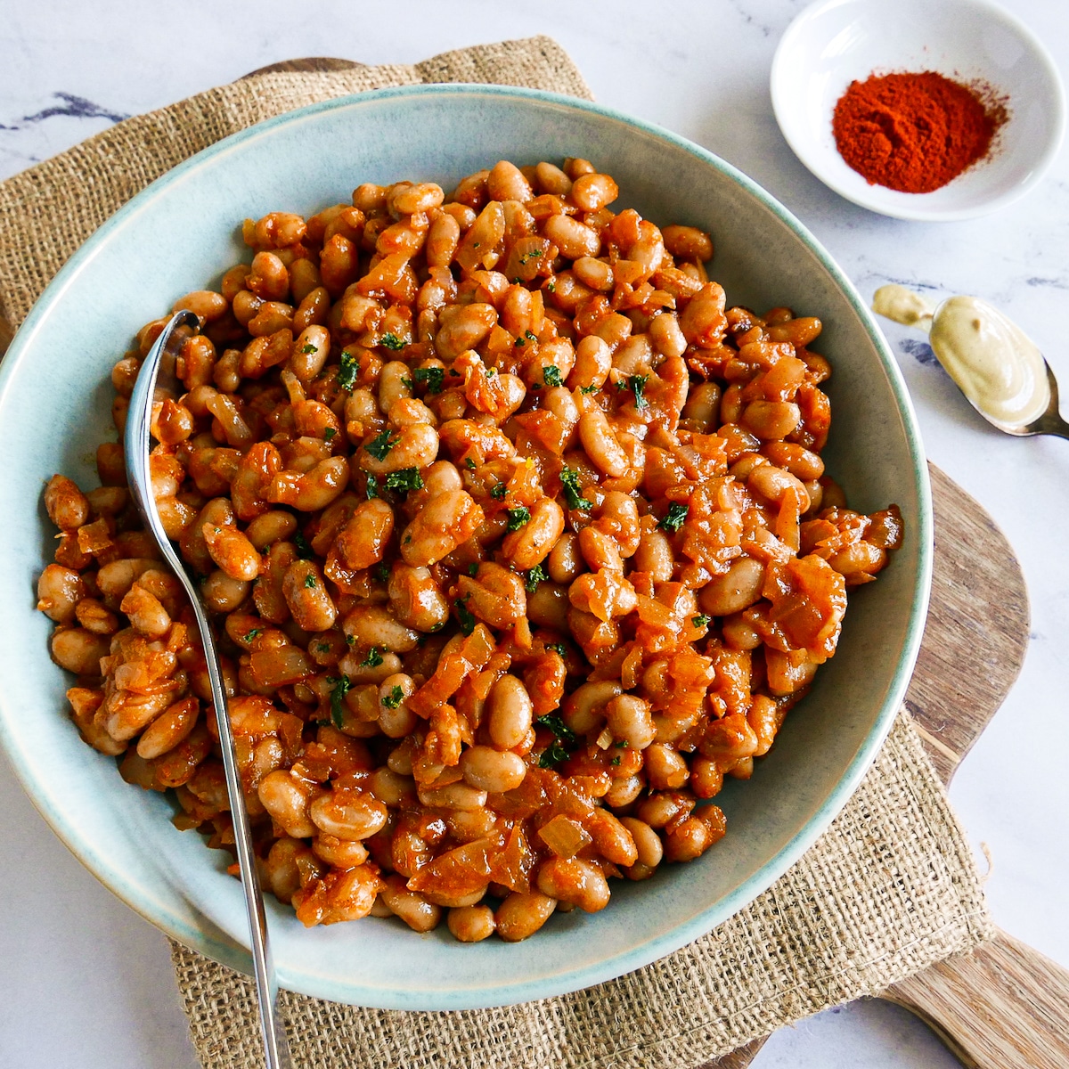 Bowl of healthy baked beans resting on a cutting board with a spoon.