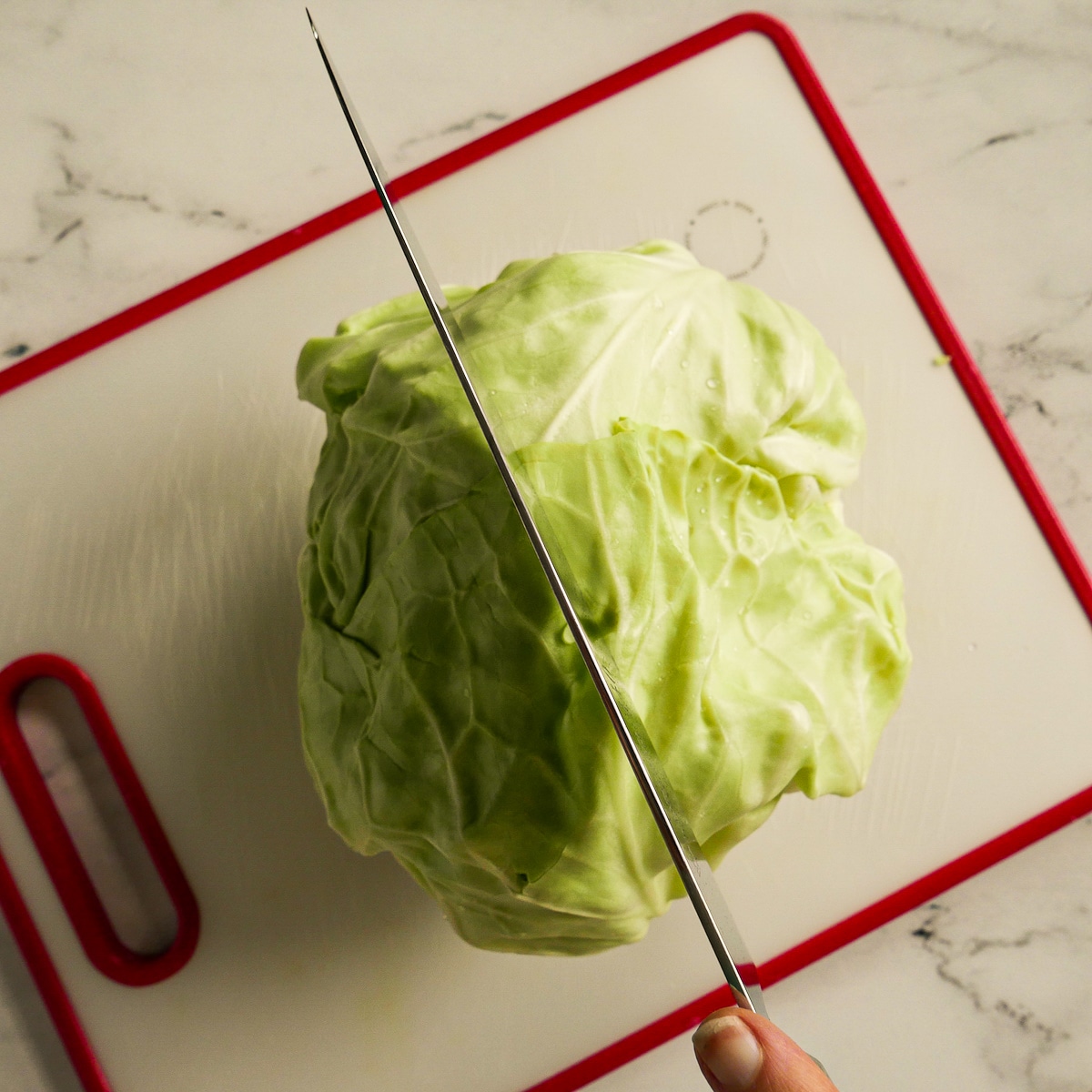 Knife slicing a green cabbage in half on a cutting board.