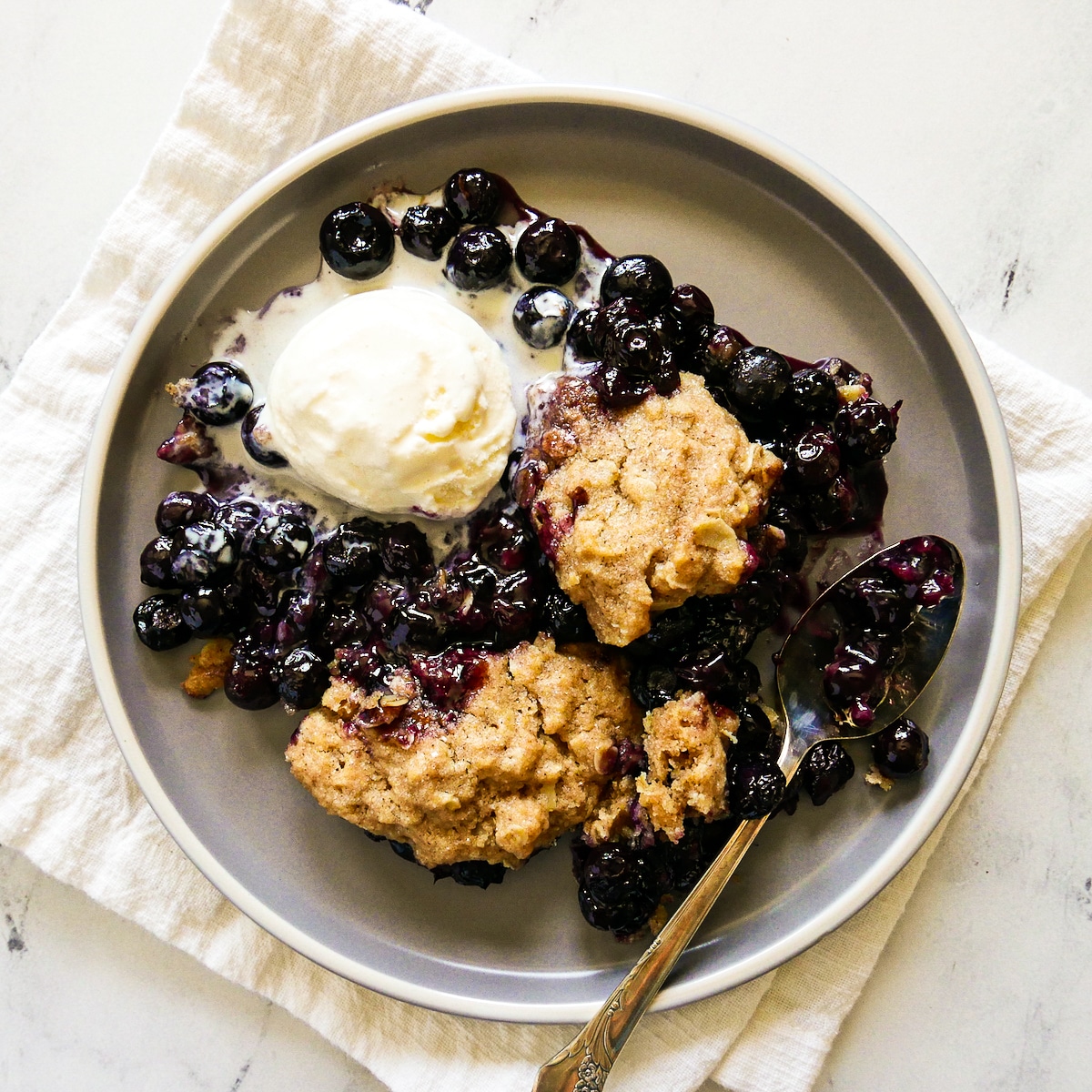 plate of blueberry cobbler with ice cream and a spoon resting on a white napkin.