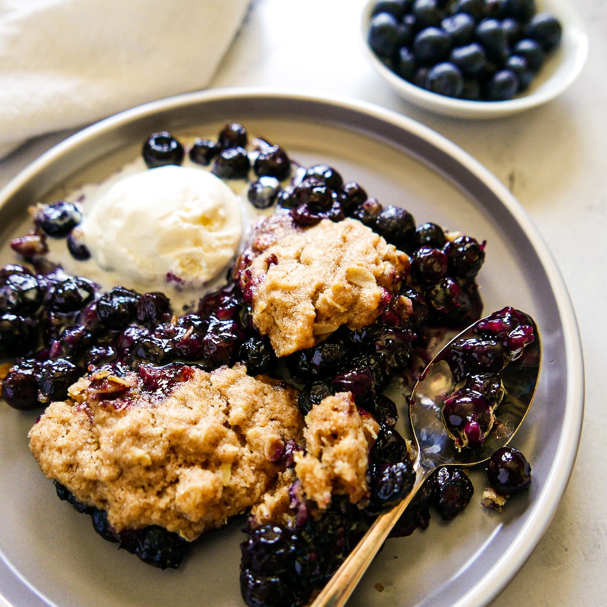 cobbler-with-ice-cream.jpg Blueberry cobbler on a plate with melting ice cream and spoon.