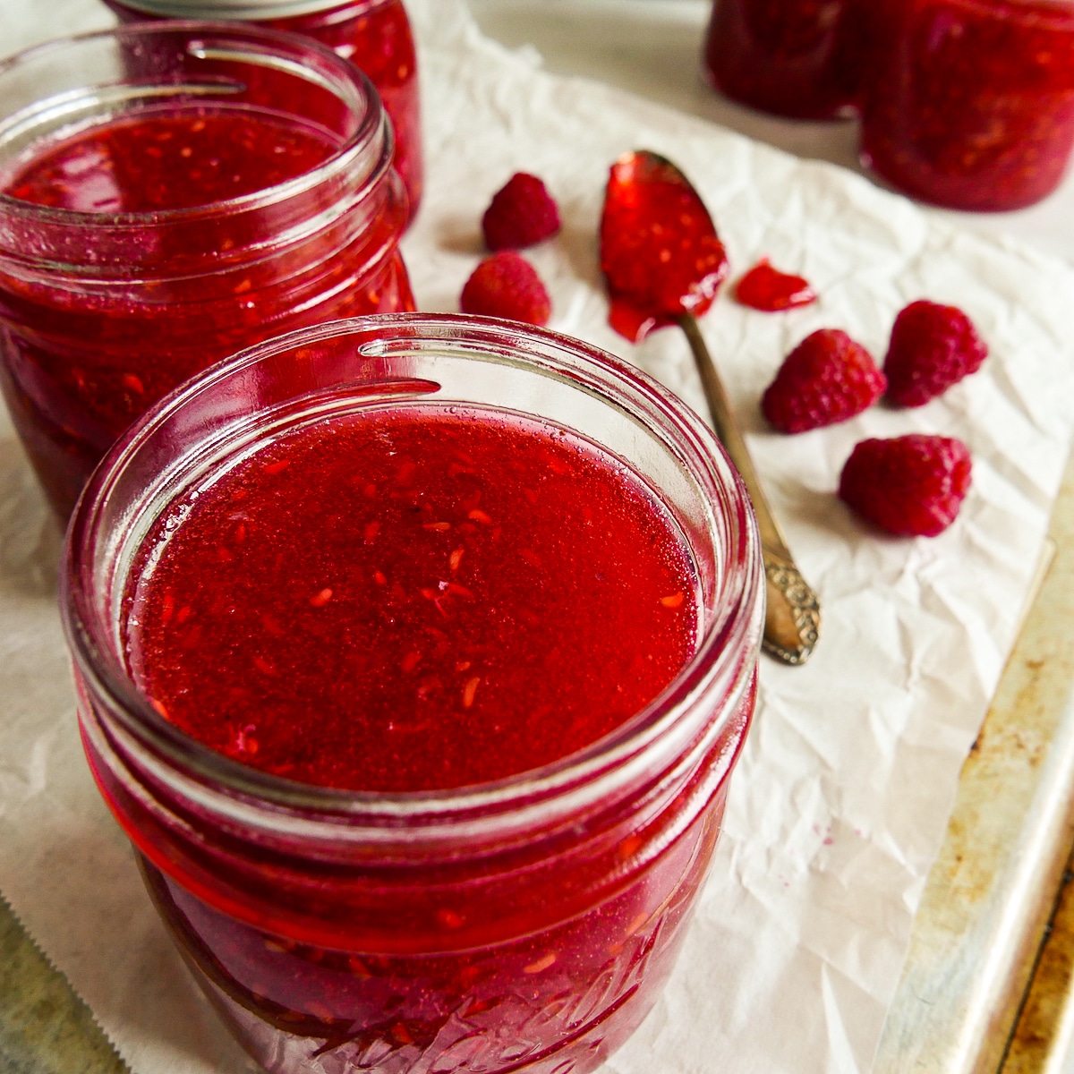 Jars of jam arranged on parchment with a spoon and raspberries.