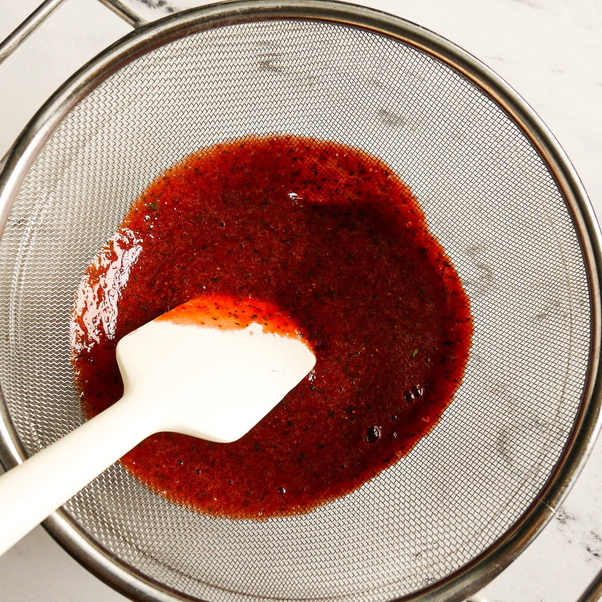 Strawberry basil mixture being pushed through a fine mesh strainer.