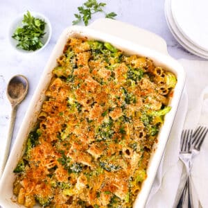 Broccoli pasta bake with plates, parsley, serving spoon, and a white napkin.