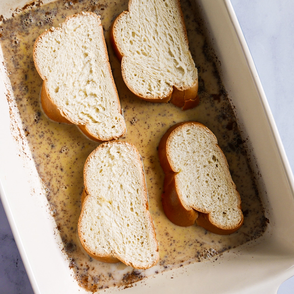 Four slices of challah bread soaking in custard bath in baking dish.