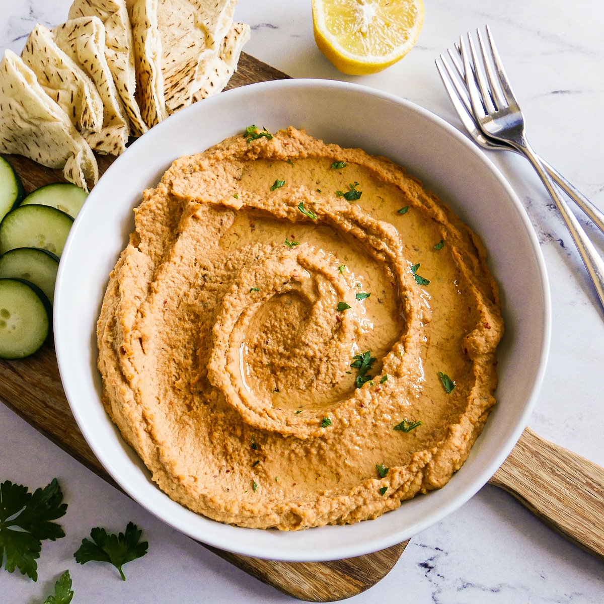 Bowl of hummus with sliced cucumber, pita, and forks.