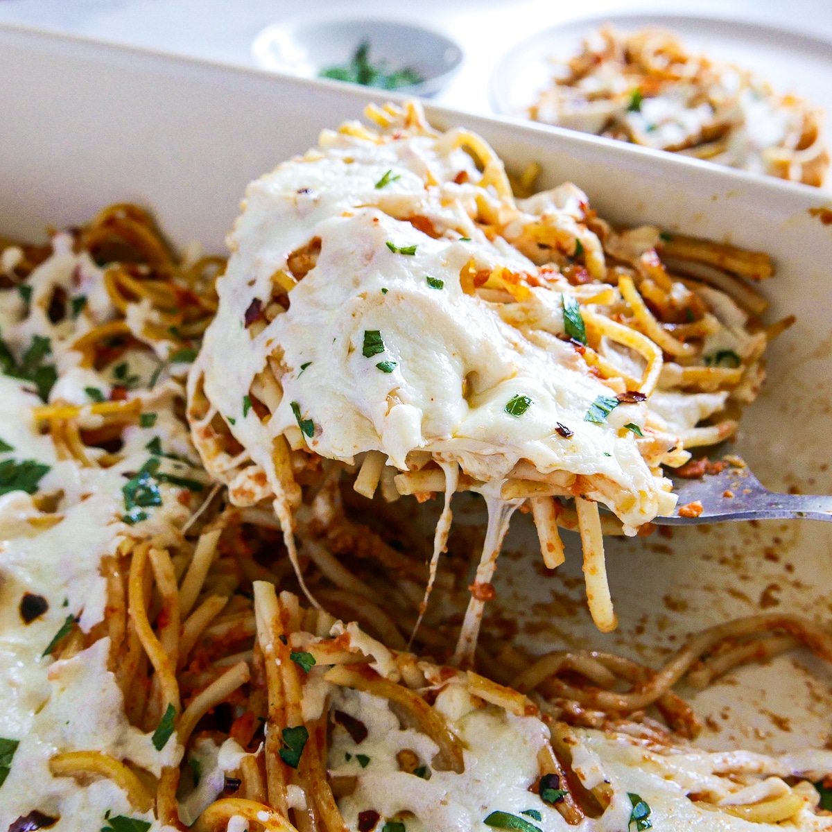 Gooey and cheesy piece of leftover spaghetti casserole being lifted out of pan.