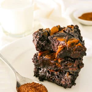 vegan pumpkin brownies stacked on a white plate with a spoon of cinnamon.