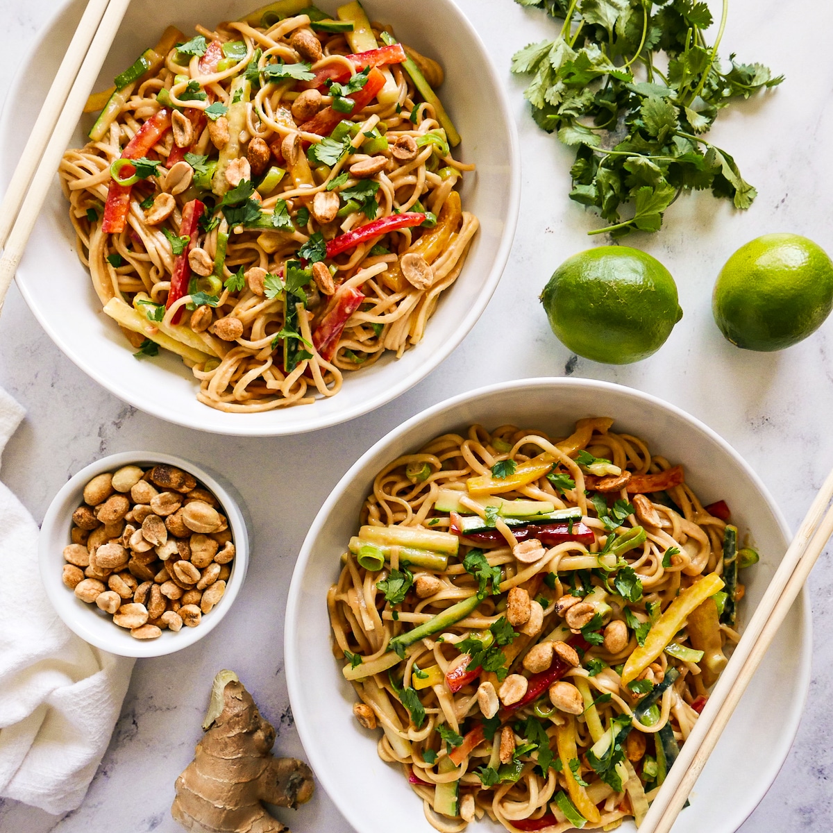 Two bowls of asian peanut noodles with peppers, cucumber, scallions, cilantro, and peanuts.