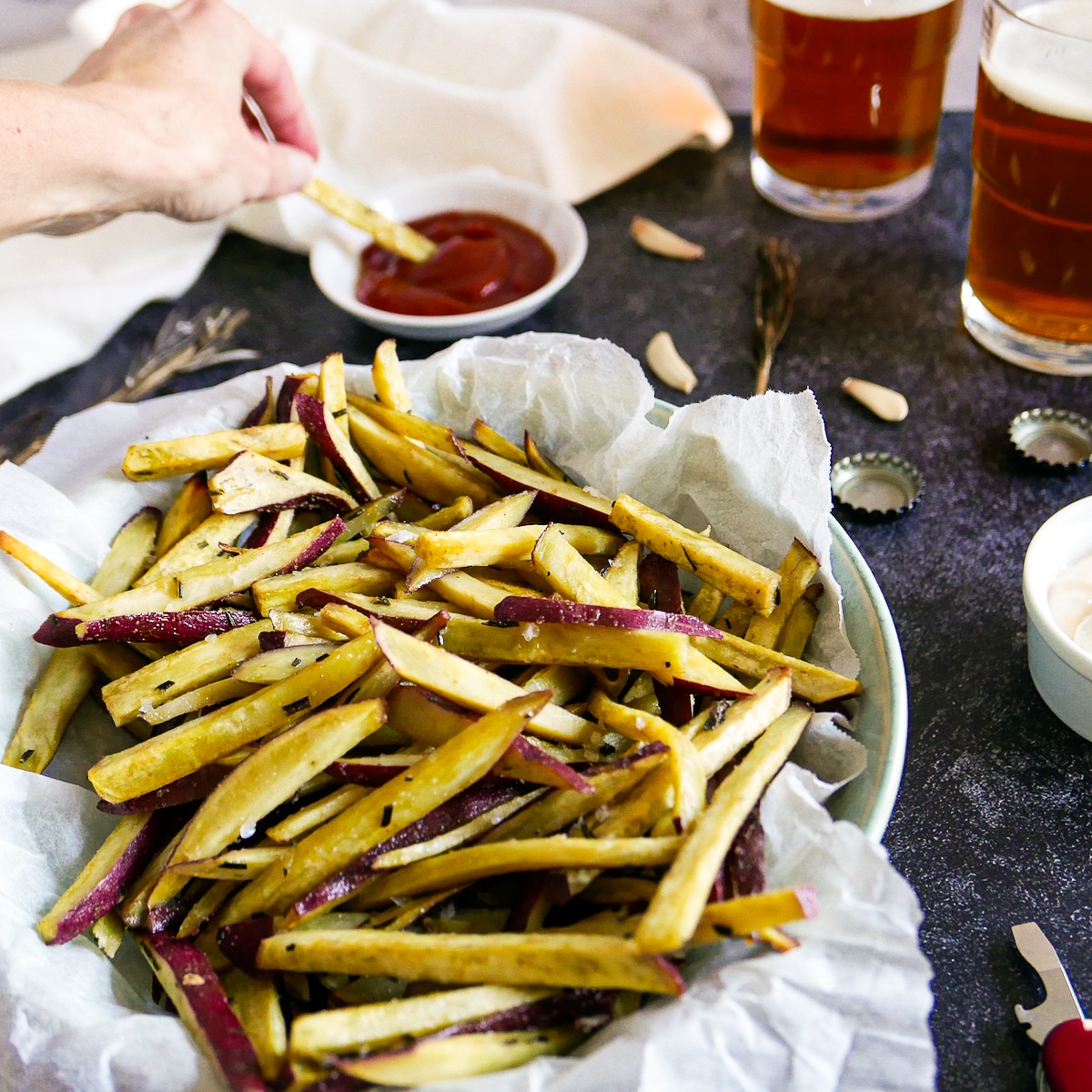 Hand dipping fries into ketchup with two glasses of beer in the background.