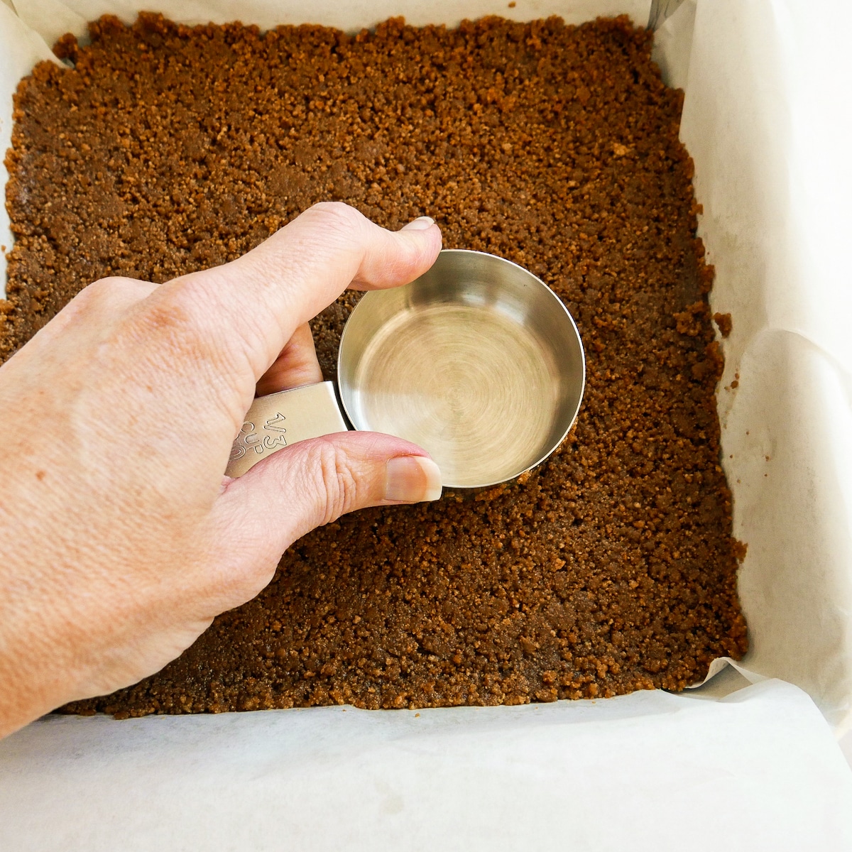 Pressing gingersnap crust into square pan using a measuring cup.