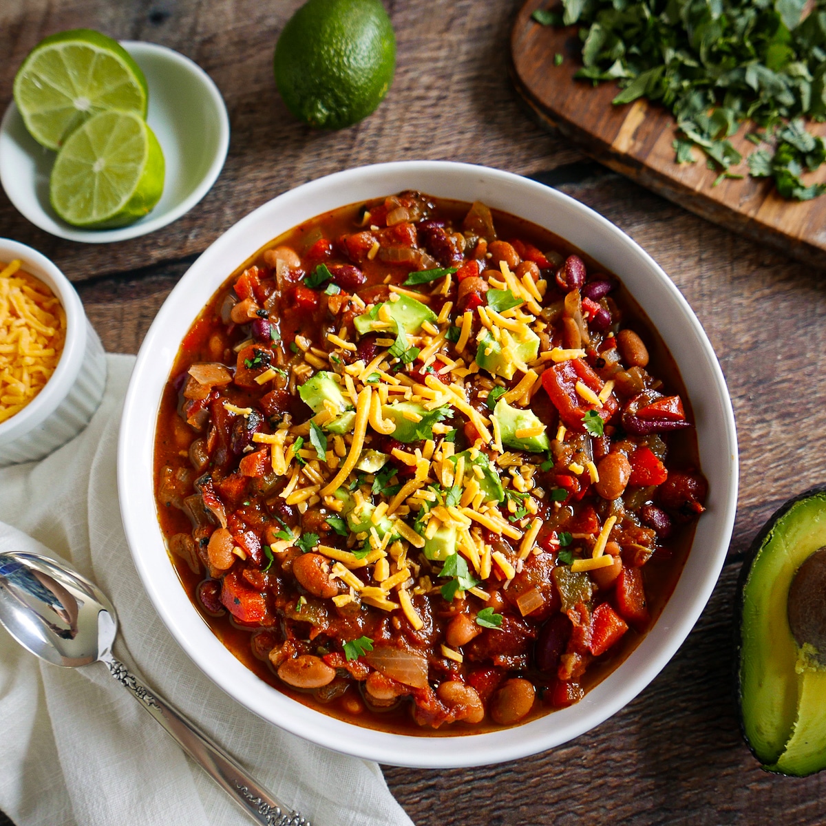 Bowl of vegetarian chili with spoon, avocado, cheese, cilantro, and lime.