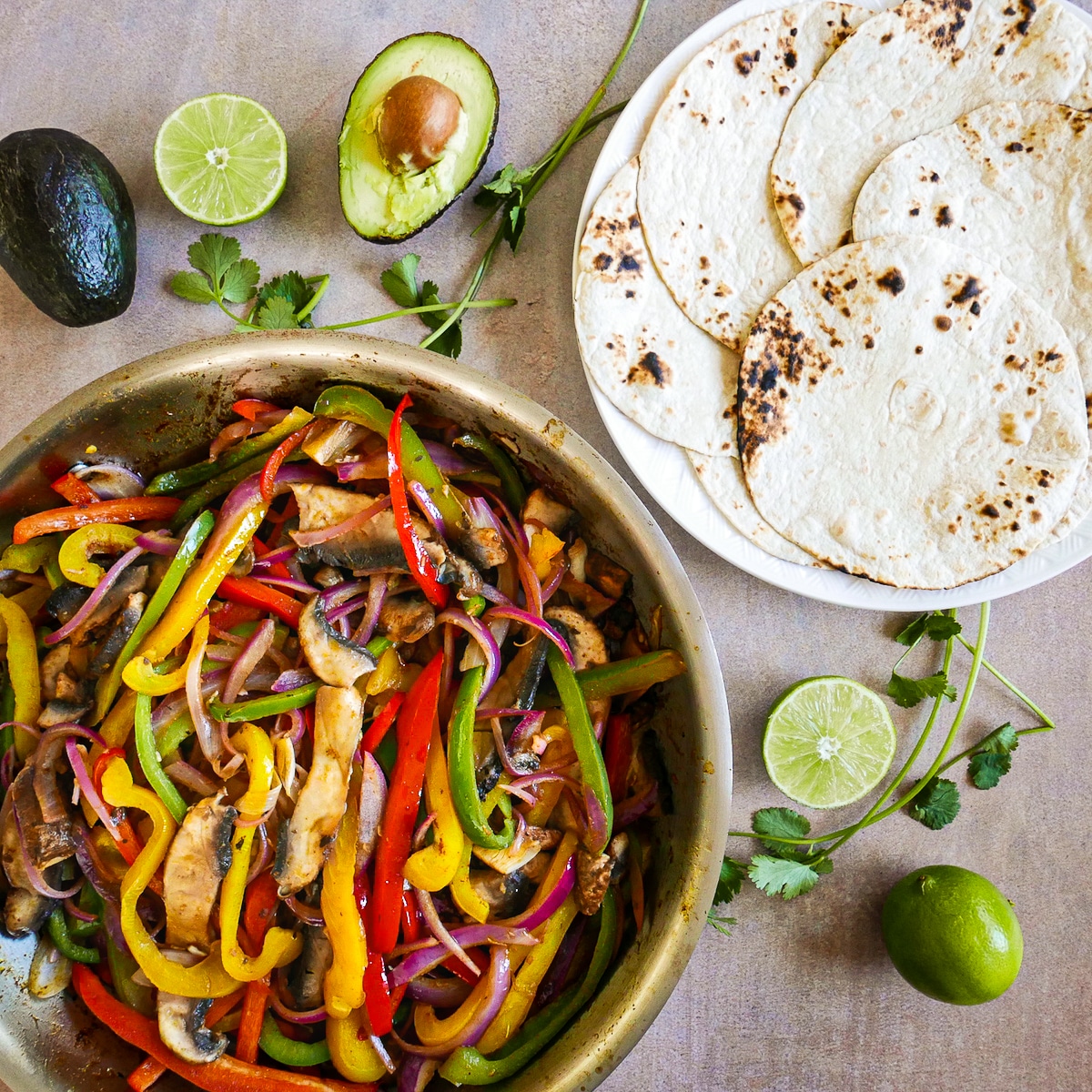 prepped-fajitas.jpg cooked veggies in skillet and flour tortillas on a plate with lime, avocado, and cilantro.