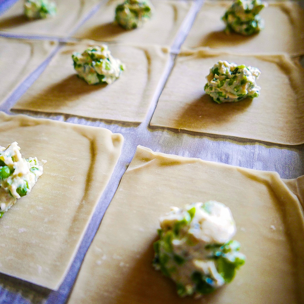 ravioli-filling-on-wrappers.jpg wonton wrappers being prepped with ricotta filling on a baking sheet.
