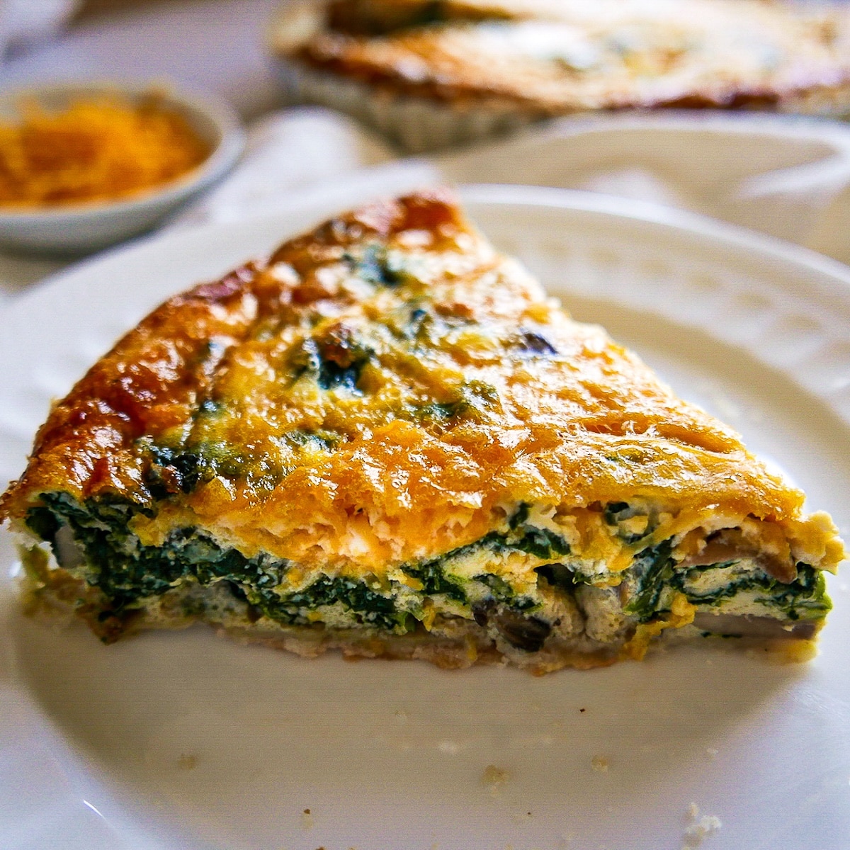 slice-of-quiche.jpg Slice of spinach quiche on a white plate with pan of quiche in background.